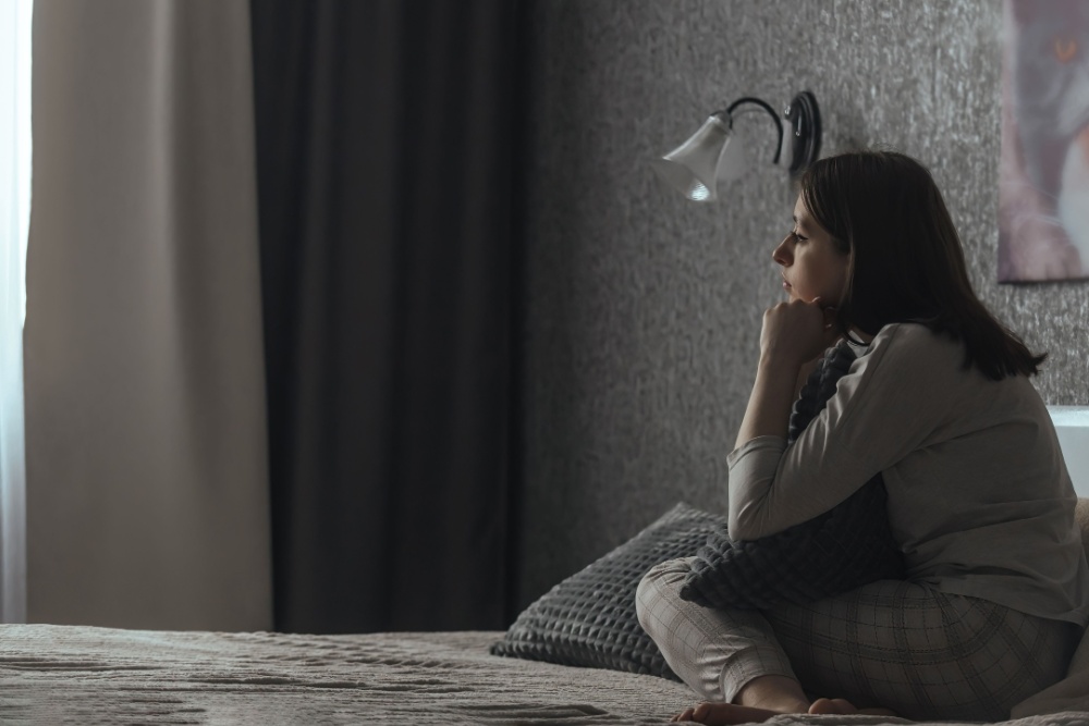 A woman sits in a dark bedroom looking sad.